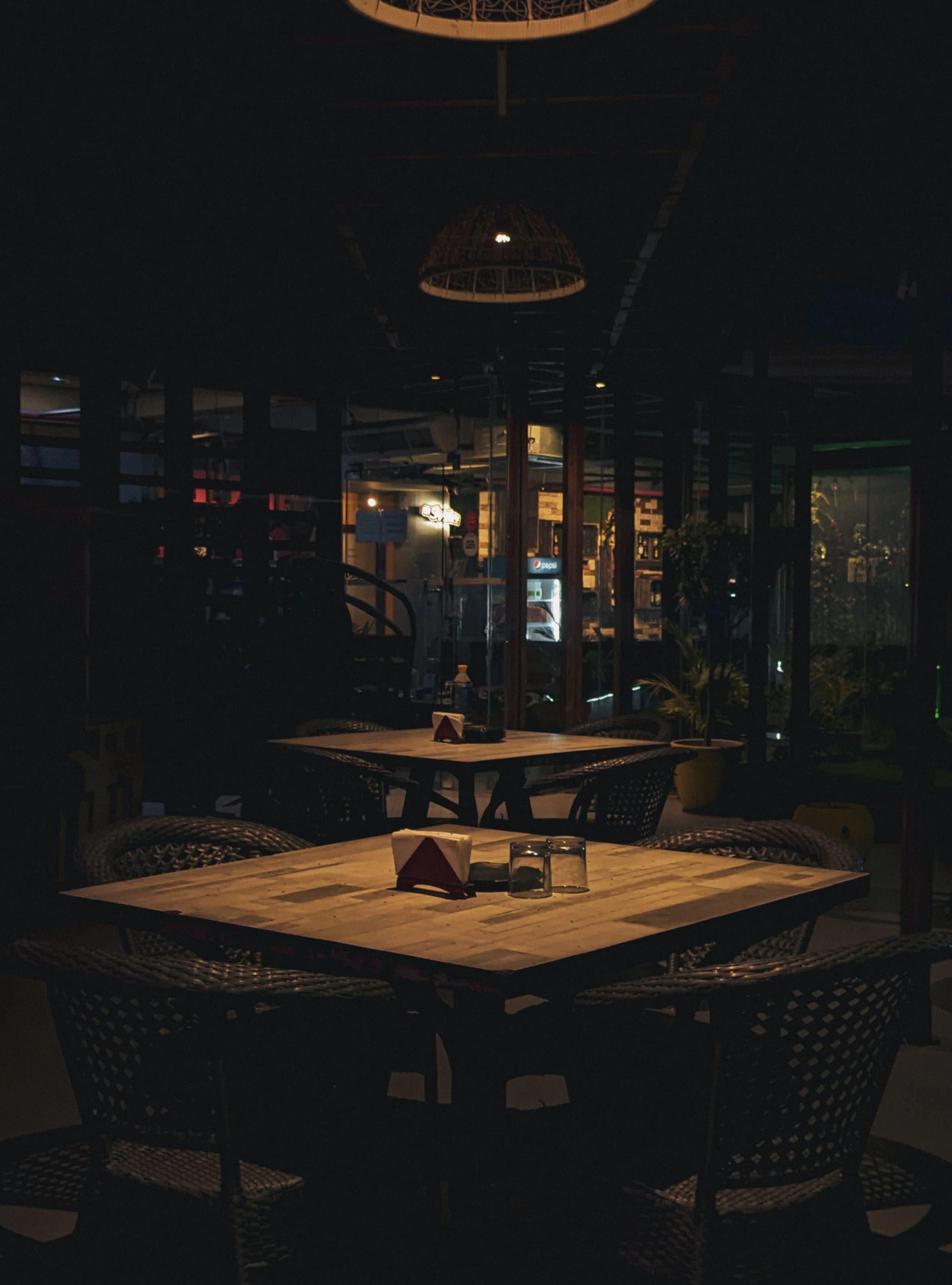 An empty four-top under a low woven lamp in a dark restaurant, set and waiting.