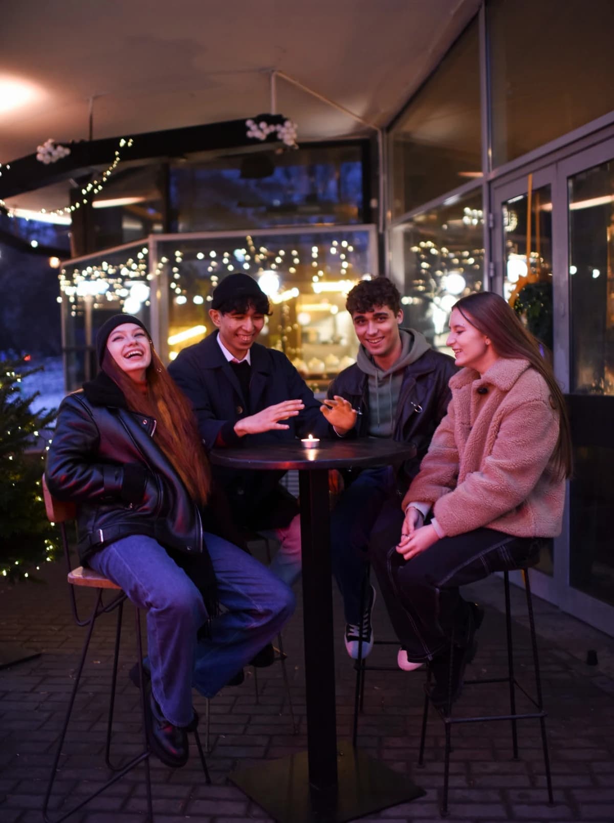 Four people laughing around a small candlelit table on a terrace strung with fairy lights.