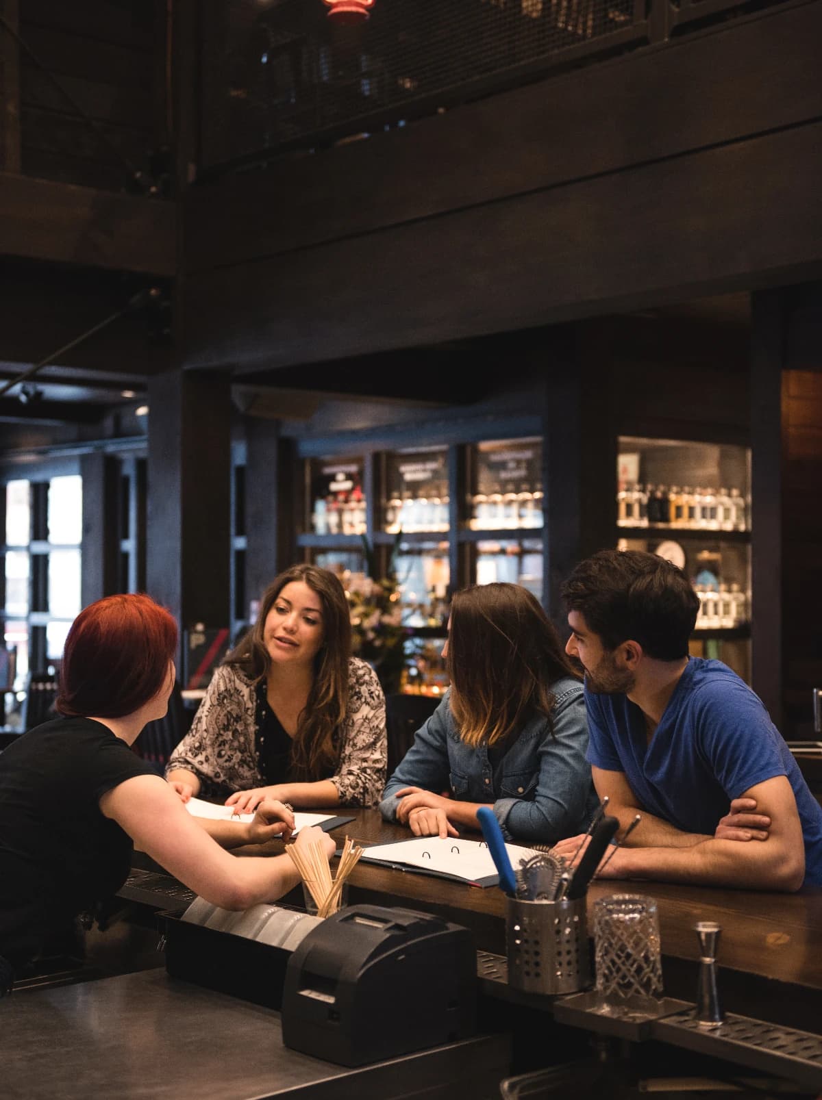 Four people talking across a bar counter while the bartender takes an order.